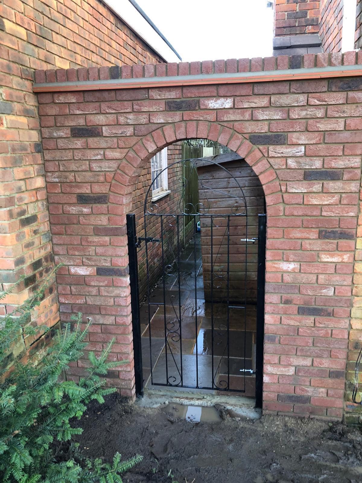 A reclaimed-brick garden wall built to an arch, with a fitted iron gate and paved threshold beneath.